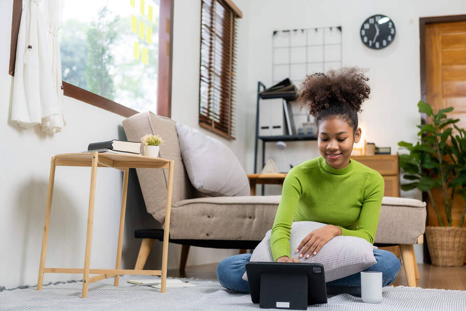 A young woman sits on the floor of her bedroom looking at her tablet with a smile